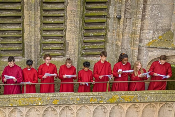 A choir in red robes, singing in a stone gallery.